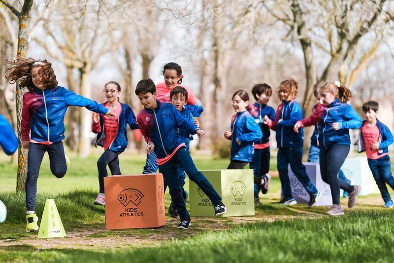 Niños jugando al aire libre, saltando cajas. Llevan chaquetas azules y rojas a juego y corren por una zona de césped.