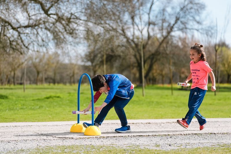 Dos niños juegan al aire libre, uno haciendo flexiones en un aro, el otro corriendo. Césped verde, árboles y un día soleado.