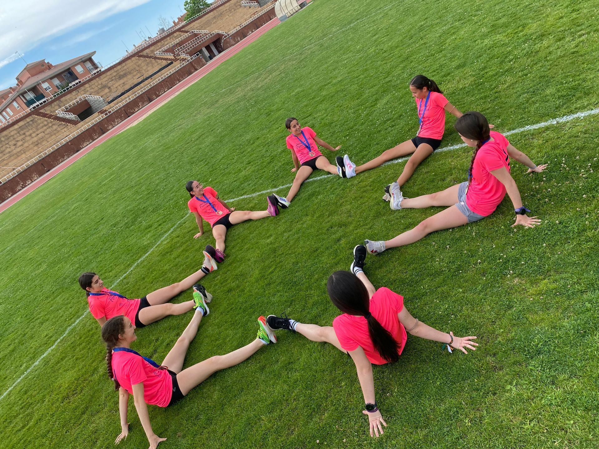 Las chicas con camisetas rosas y pantalones cortos se estiran en círculo en un campo verde, con un estadio al fondo.