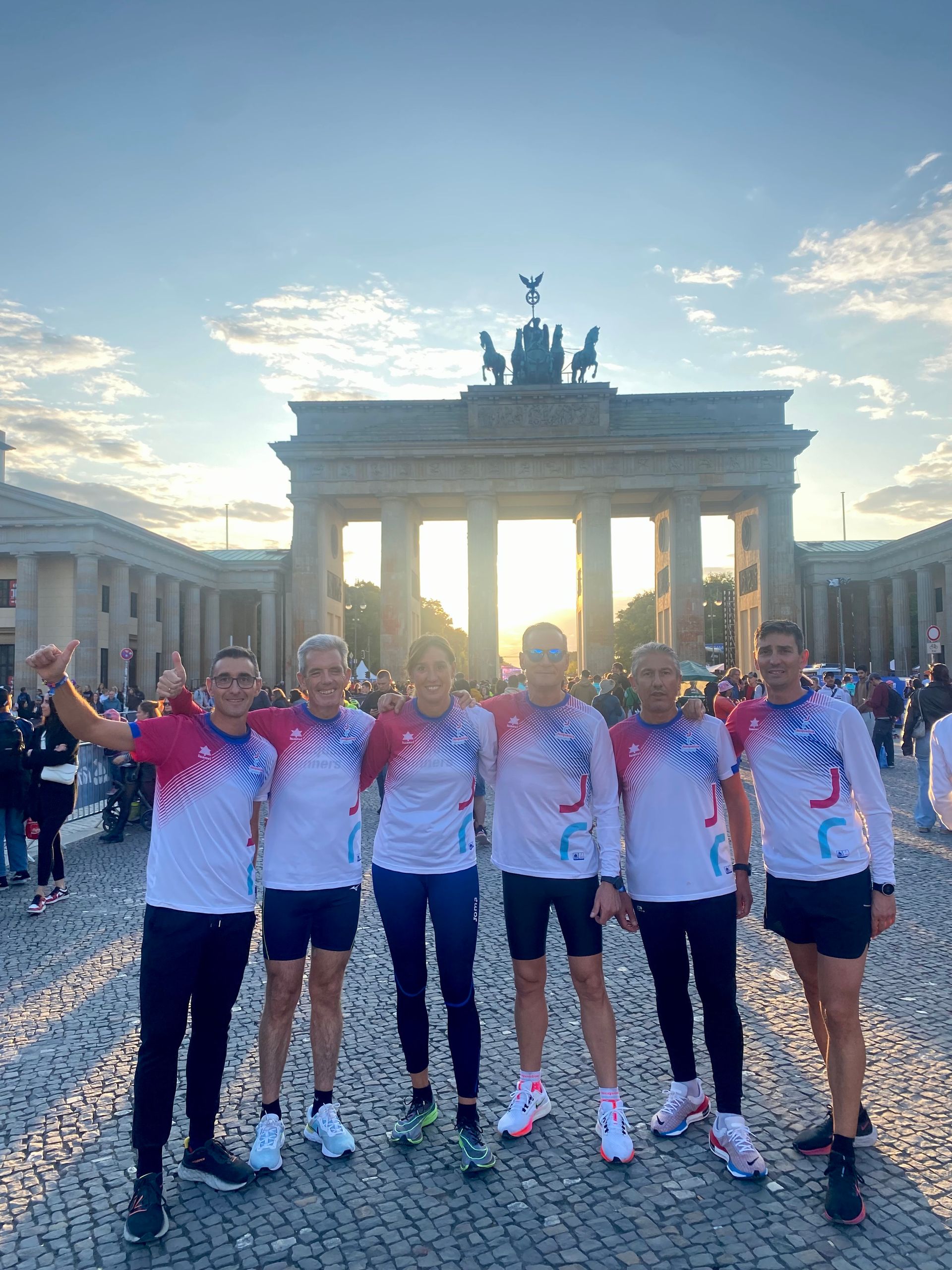 Un grupo de corredores frente a la Puerta de Brandeburgo en Berlín. Visten camisetas iguales y celebran con una sonrisa.